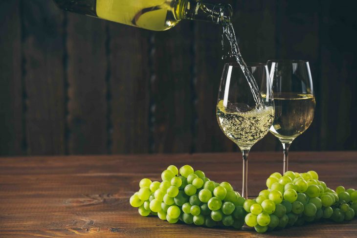 Pouring white wine from the bottle into a glass with a bunch of green grapes against wooden background with free space