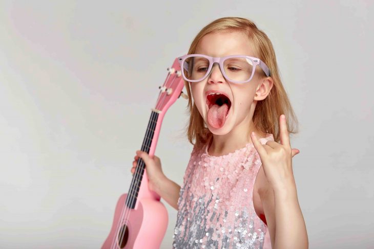 Rock and roll music concept. Cute caucasian little girl shows her tongue in a bright pink dress and glasses are playing the ukulele on a gray studio background with copy space