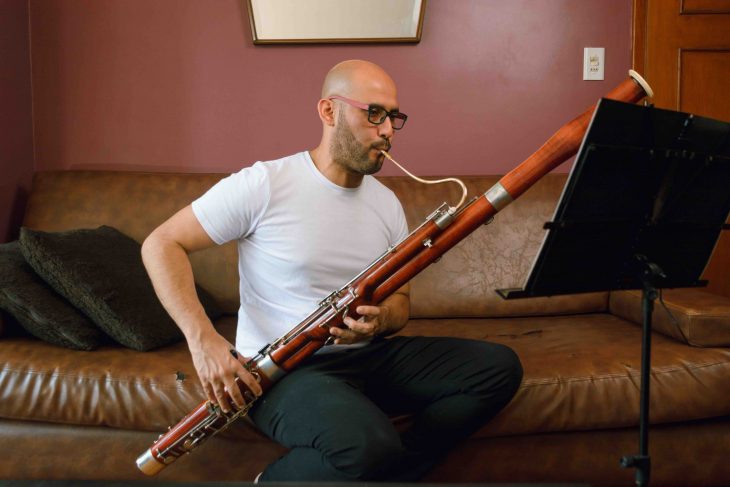 young latino man, bald with glasses and a white shirt, is sitting on a leather sofa playing the bassoon, reading and studying sheet music at home.