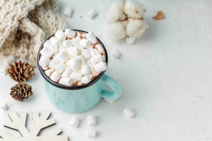 Blue metal mug of cocoa ot hot chocolate with marshmallows on the table with winter decor, branch of cotton flower, knitted blanket and wooden Christmas ornaments