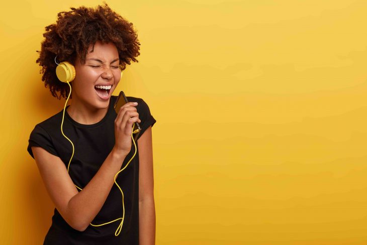 Delighted happy dark skinned emotional woman feels herself as real star, dances and sings along music, uses smart phone as microphone, modern headphones, stands over yellow studio wall, enjoys life