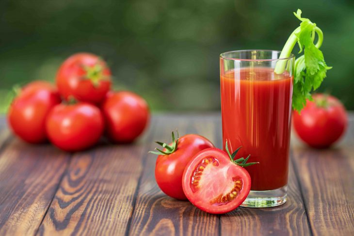 fresh tomato juice in glass with celery stick and heap of ripe vegetables on wooden table outdoors